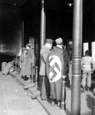 US soldiers standing at a train station. A GI with his back to the camera is wearing a Nazi flag.jpg